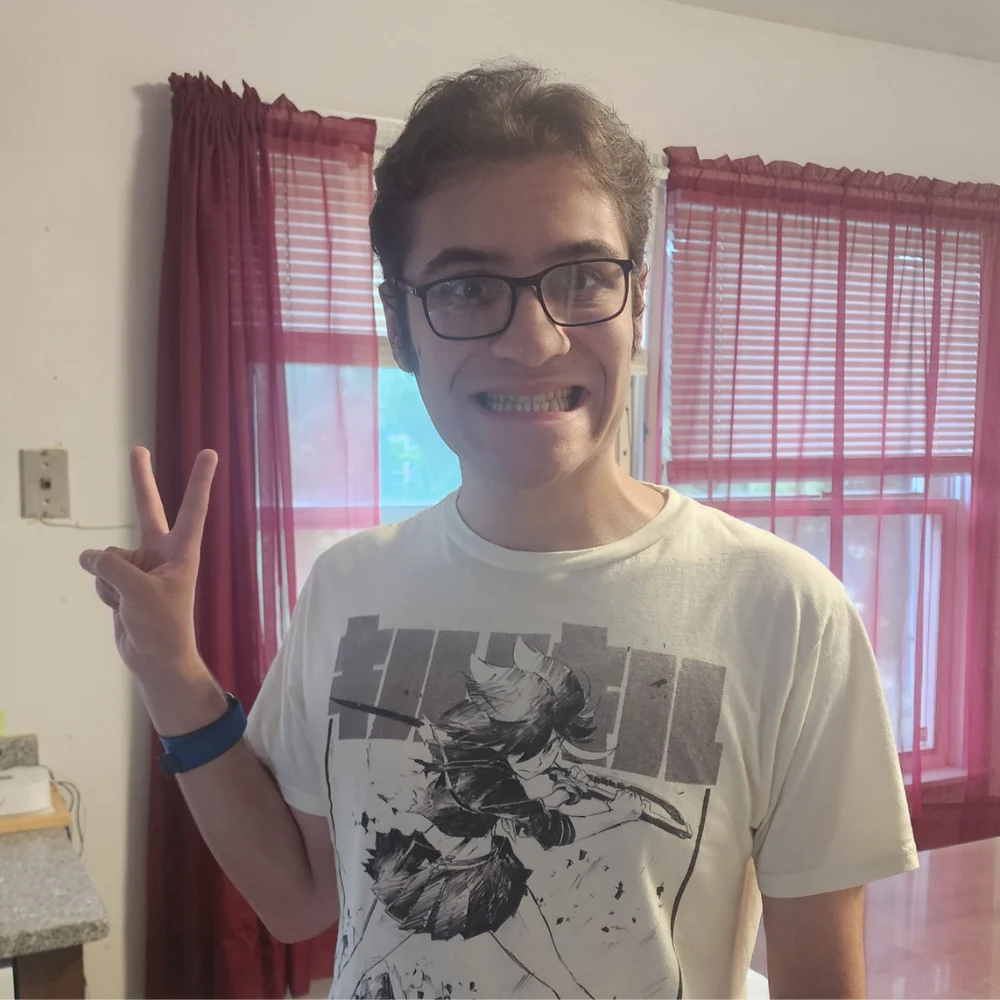 Move in Day 2 A young man wearing glasses and a graphic anime t-shirt smiles cheerfully while making a peace sign gesture with his hand indoors in front of a window with red sheer curtains.