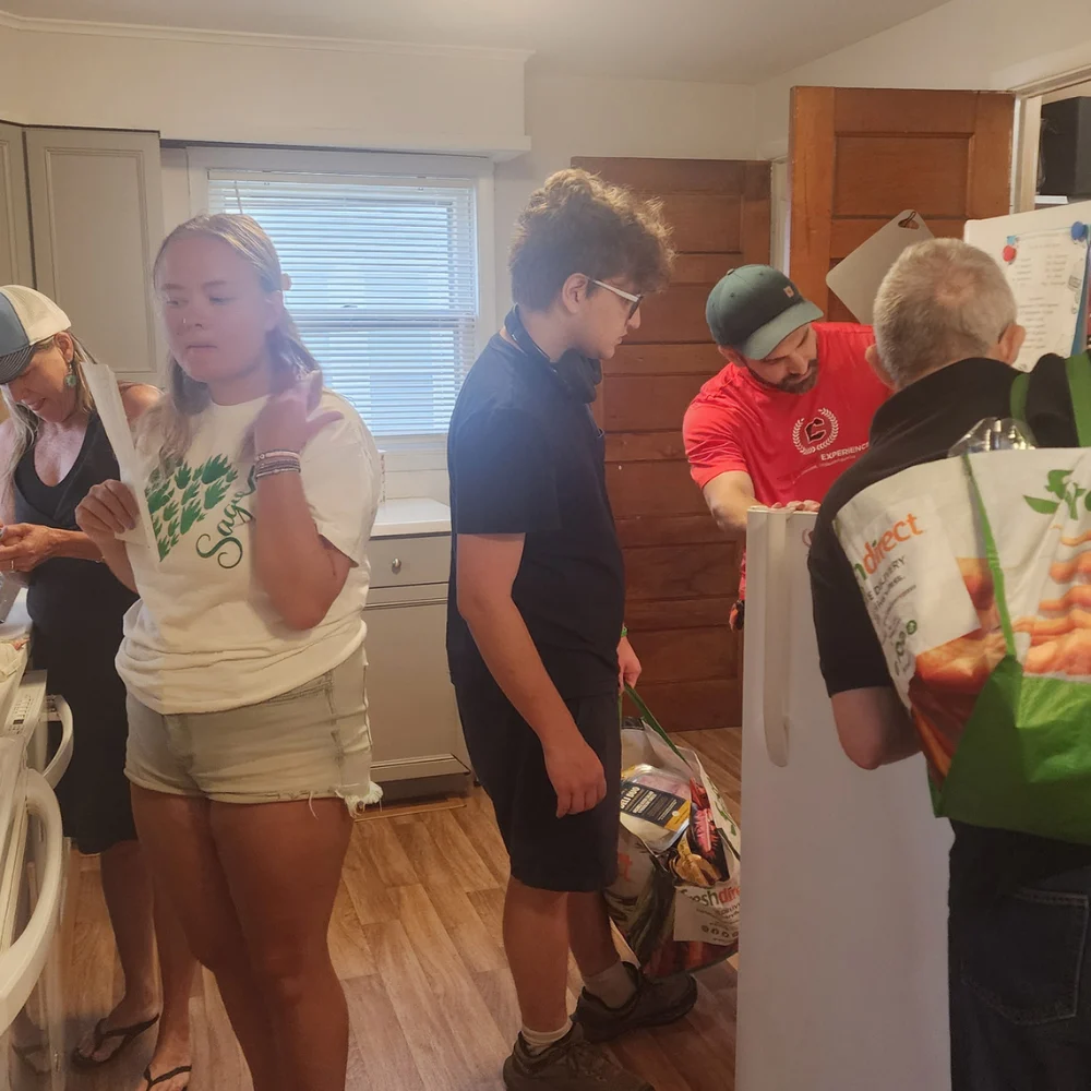 Move in day 3 A group of people inside a brightly lit kitchen unpacking groceries and moving items. A young woman in a white graphic shirt holds a piece of paper, a young man carries a grocery tote bag, and others organize items near the refrigerator.
