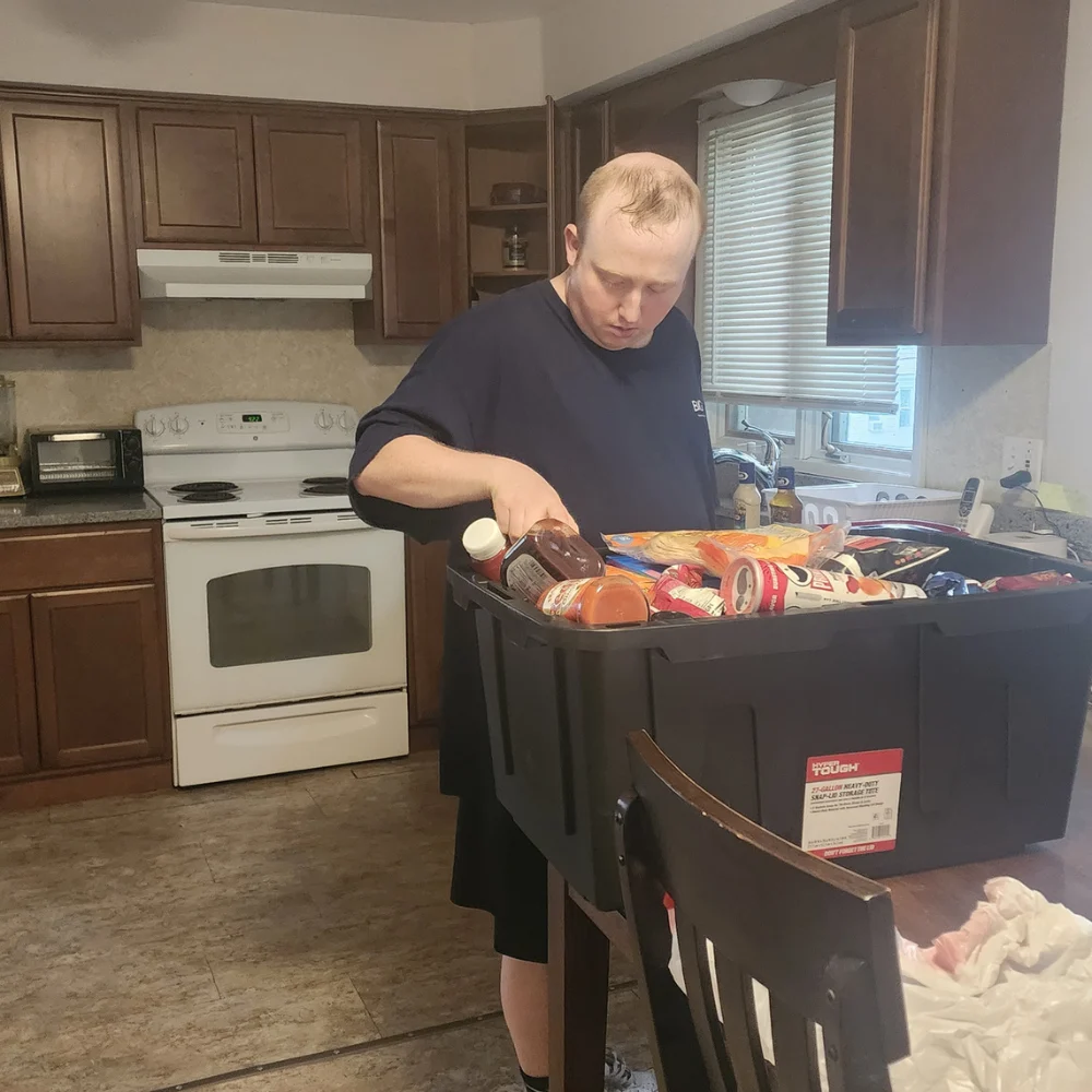 Move in day 4 A man standing in a kitchen unpacks groceries from a large black plastic storage tote placed on a dining table. The kitchen background features brown wooden cabinets, a white stove, and window blinds.