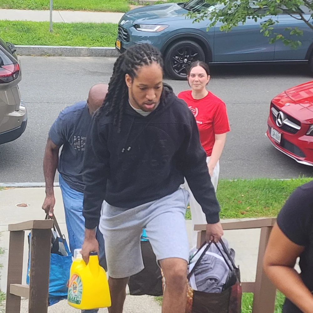 Move in day 5 A young man wears a black hoodie and grey shorts while carrying a yellow detergent bottle and tote bags up outdoor stairs. Other individuals follow behind him carrying bags from parked vehicles on the street.