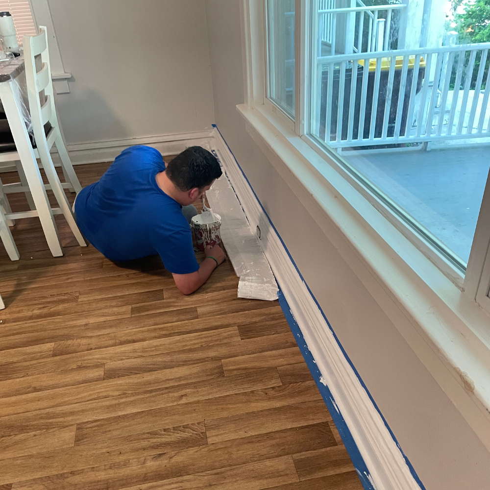 Painting Trim 1 A man wearing a royal blue t-shirt leans low over a light wood laminate floor, using a small brush and paint container to carefully apply white paint to the baseboard trim along a large window wall lined with blue painter's tape.