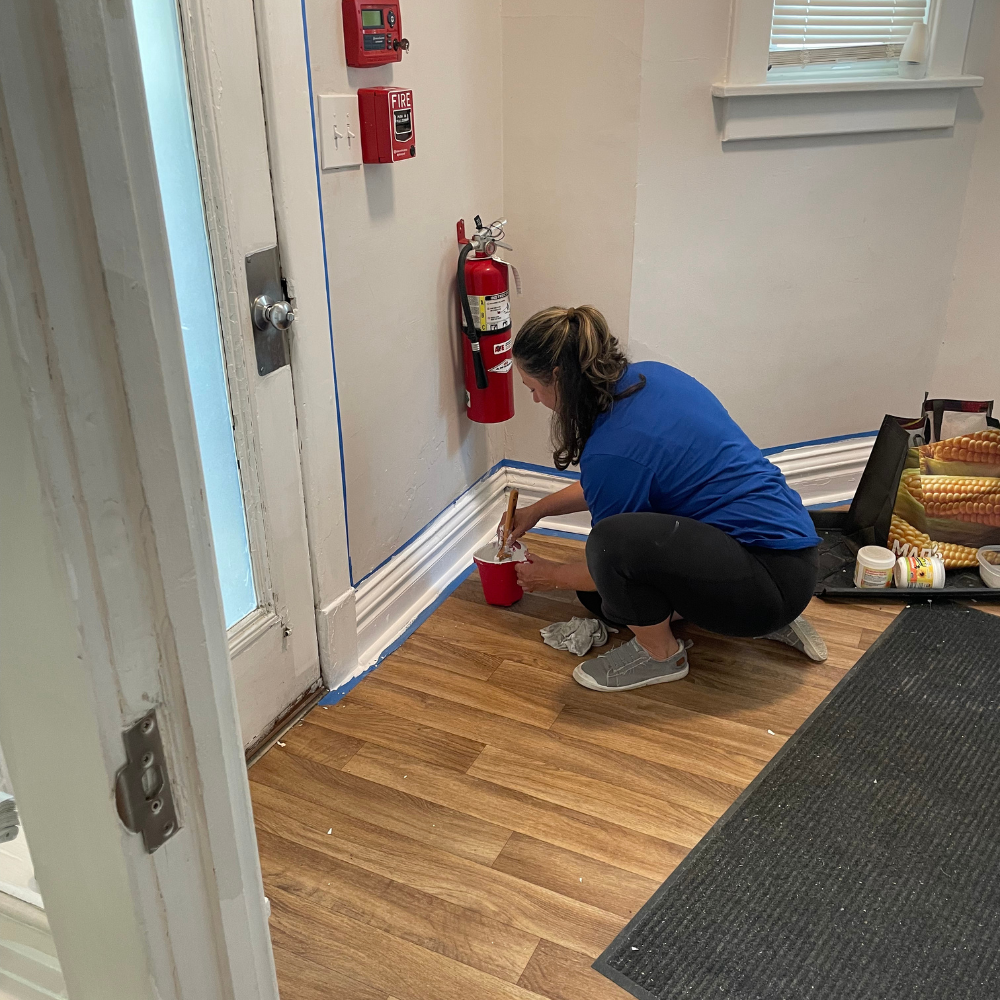 Painting Trim 2 A woman with her hair in a ponytail squats on a wood floor near a front door and a wall-mounted red fire extinguisher. She uses a paintbrush and a red plastic cup filled with white paint to touch up white baseboard trim prepped with painter's tape.