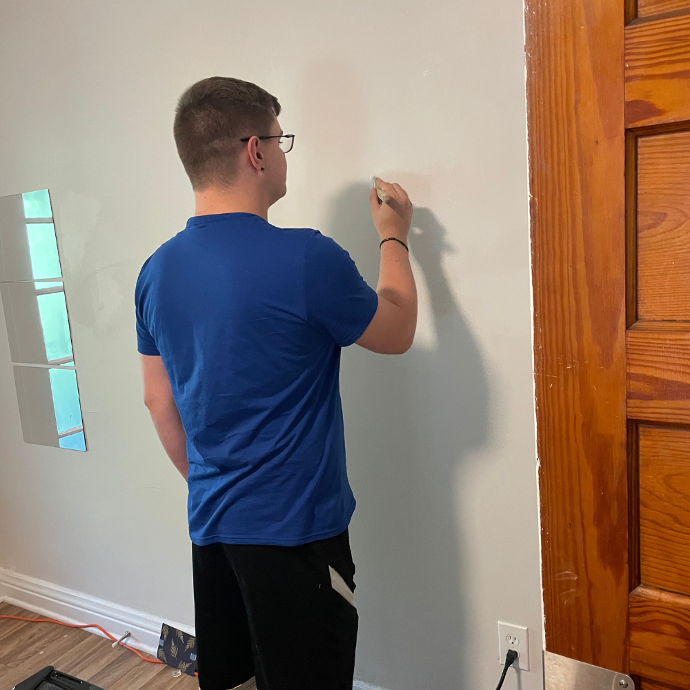Painting Wall 1 View from behind of a young man wearing glasses, a blue t-shirt, and black shorts as he uses a small paintbrush to carefully touch up a light grey interior wall next to a natural wood door frame.