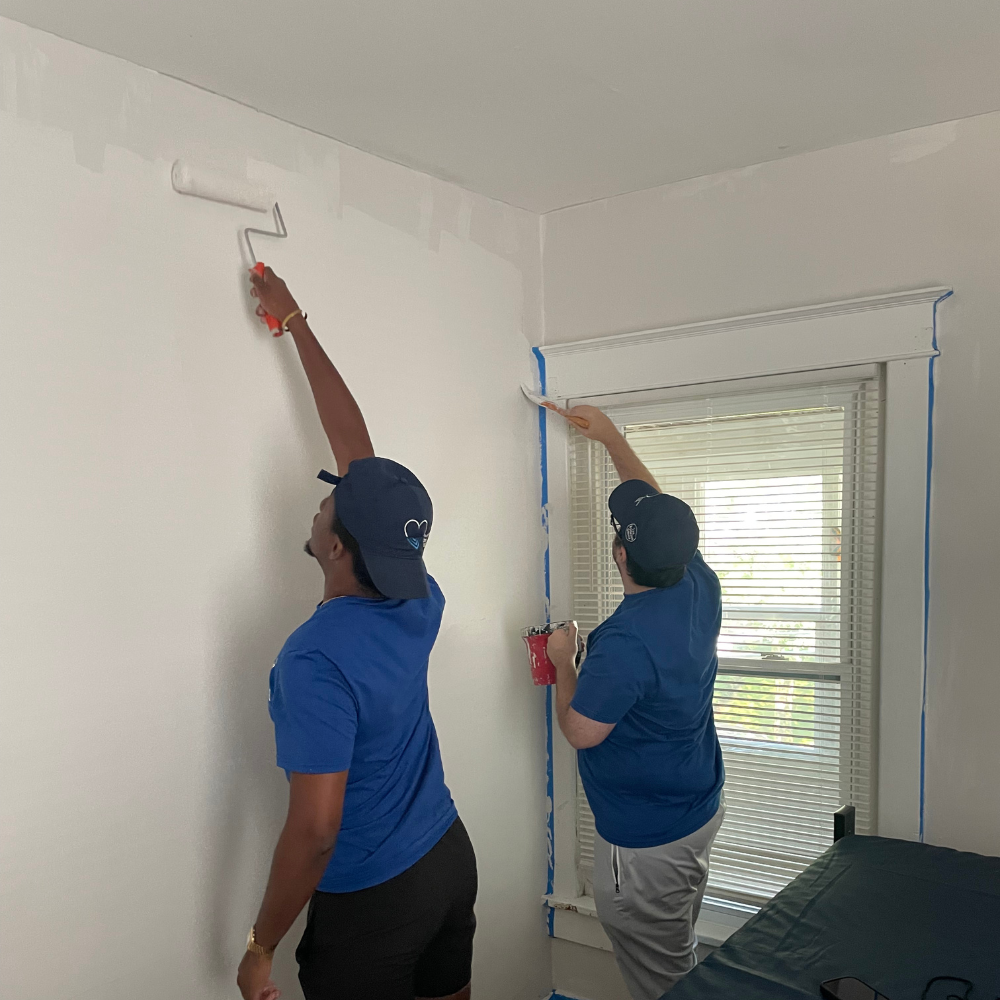 Painting Walls Two male volunteers wearing royal blue t-shirts and dark baseball caps work together indoors. The person in the foreground uses a paint roller to reach high on a white wall, while the person in the background uses a brush to paint the window trim framed in blue painter's tape.