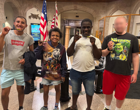 Four people standing in a row inside a museum or historic building, each holding up a small yellow pen or souvenir. Two American flags and a large historic cannon are visible in the background.