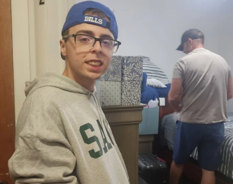 A young man wearing glasses, a blue "BILLS" baseball cap turned backward, and a grey hoodie smiles at the camera inside a bedroom. In the background, another person works near a bed surrounded by stacked decorative storage boxes, suitcases, and pillows.