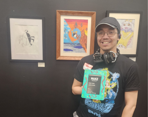 A smiling person stands in front of a dark gallery wall featuring three framed artworks. They wear a black Dragon Ball Z graphic t-shirt, a red name tag reading "Tyler", gray camouflage cargo pants, white sneakers, a dark baseball cap, glasses, and black headphones around their neck. In one hand, they hold a water bottle, and with the other, they display a bright turquoise painted picture frame with green and orange accent designs. Behind them on the wall are three framed pieces: a black-and-white minimalist insect drawing on the left, a colorful abstract landscape painting in a wooden frame in the middle, and an illustration of an anime-style character in a black frame on the right.