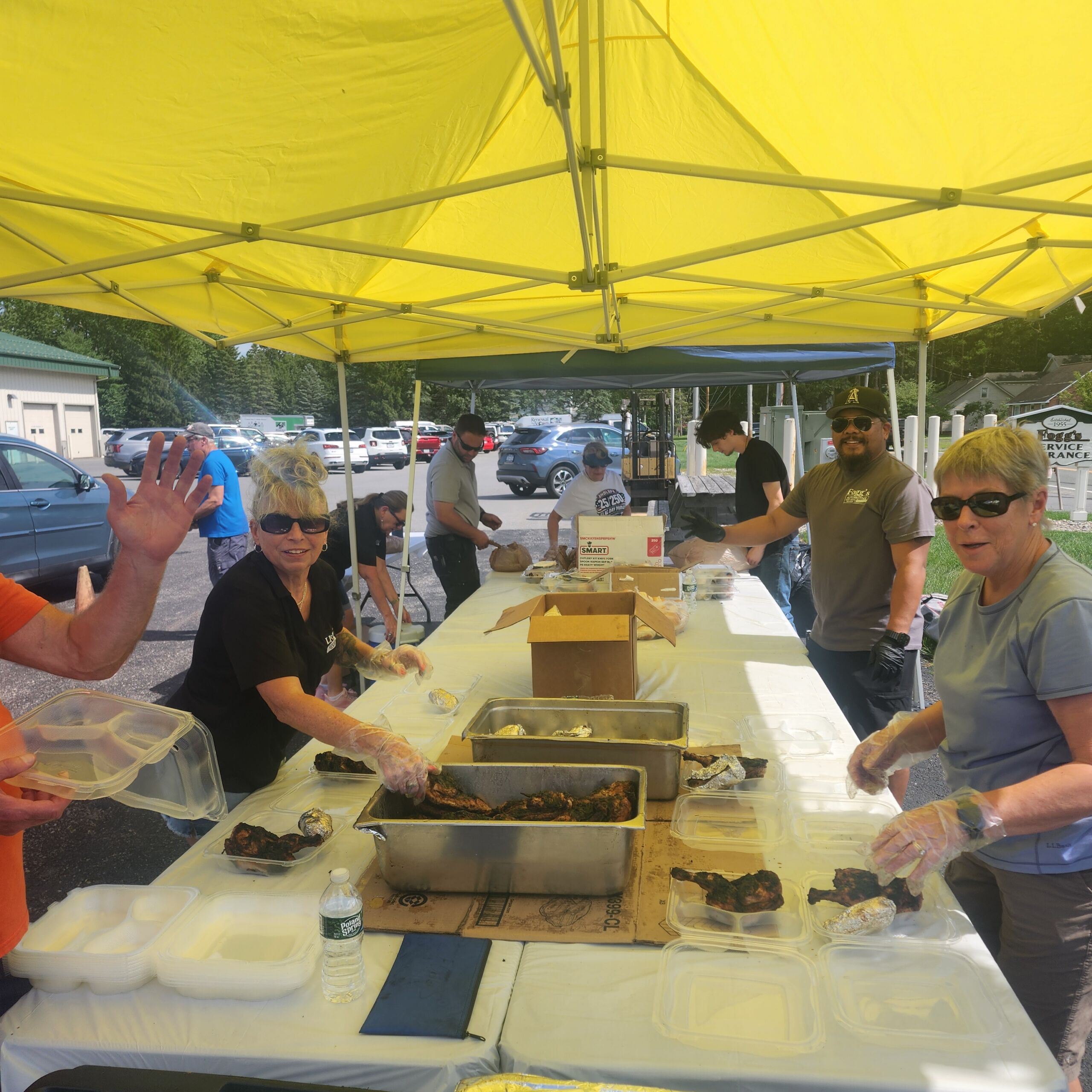 Volunteers Volunteers wearing gloves package fresh Brooks BBQ chicken into plastic clam-shell containers at long tables set up under a bright yellow canopy tent outdoors.