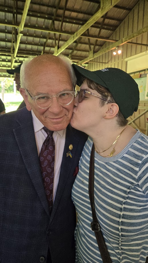 Tonko Brain Injury Picnic A person wearing a green baseball cap, glasses, and a blue-and-white striped shirt giving a cheek kiss to an older, smiling man wearing clear-framed glasses, a dark plaid suit jacket, and a paisley tie under an outdoor pavilion.