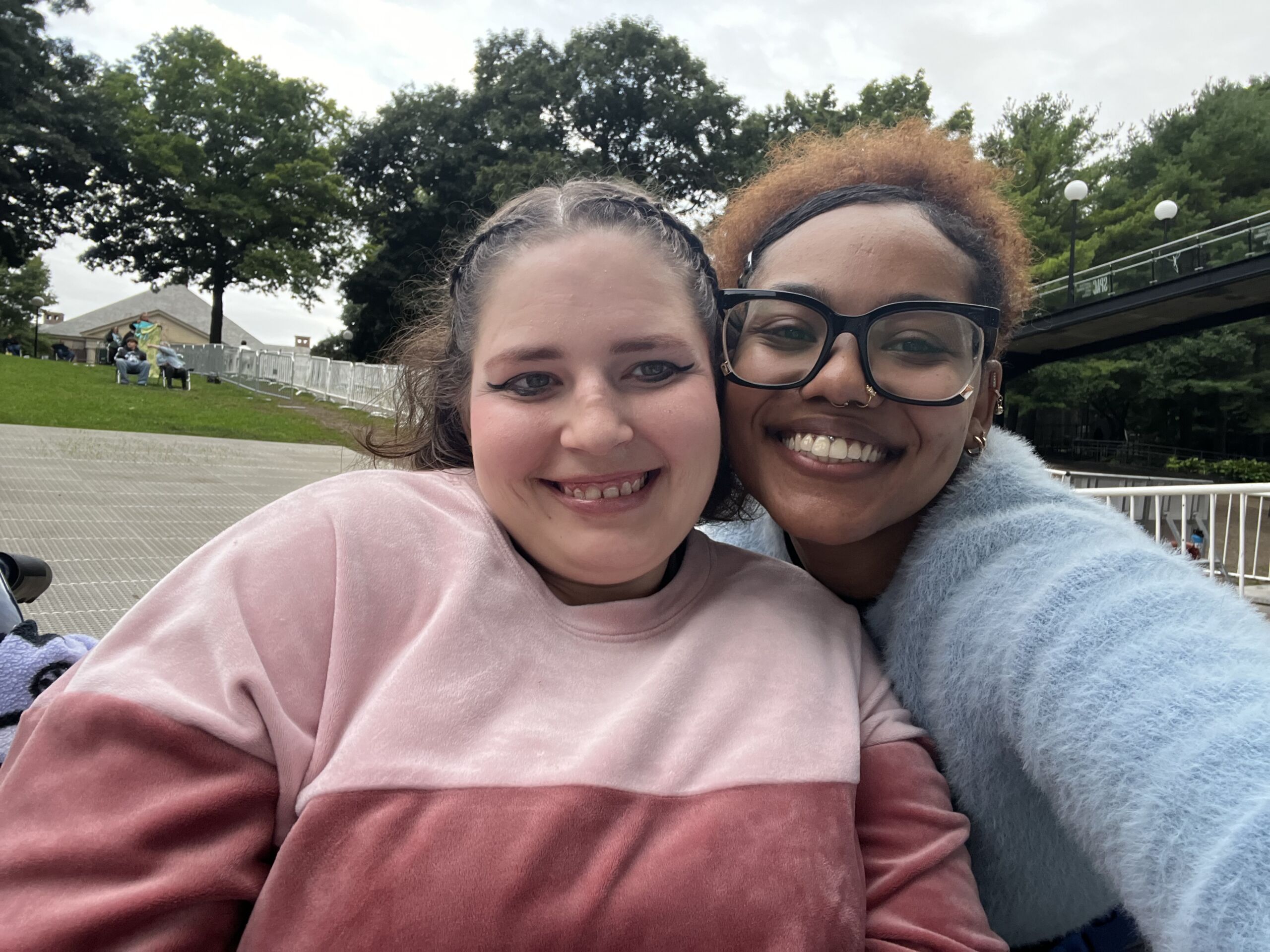 Pitbull Concert 1 Daytime outdoor selfie of two friends smiling closely together. The person on the left has dark hair styled in french braids and is wearing winged eyeliner and a color-blocked pink sweatshirt. The person on the right has auburn hair tied back, black rimmed glasses, a nose ring, and a fuzzy light blue sweater. The background shows a grassy park area with green trees, white barricades, and an overcast sky.
