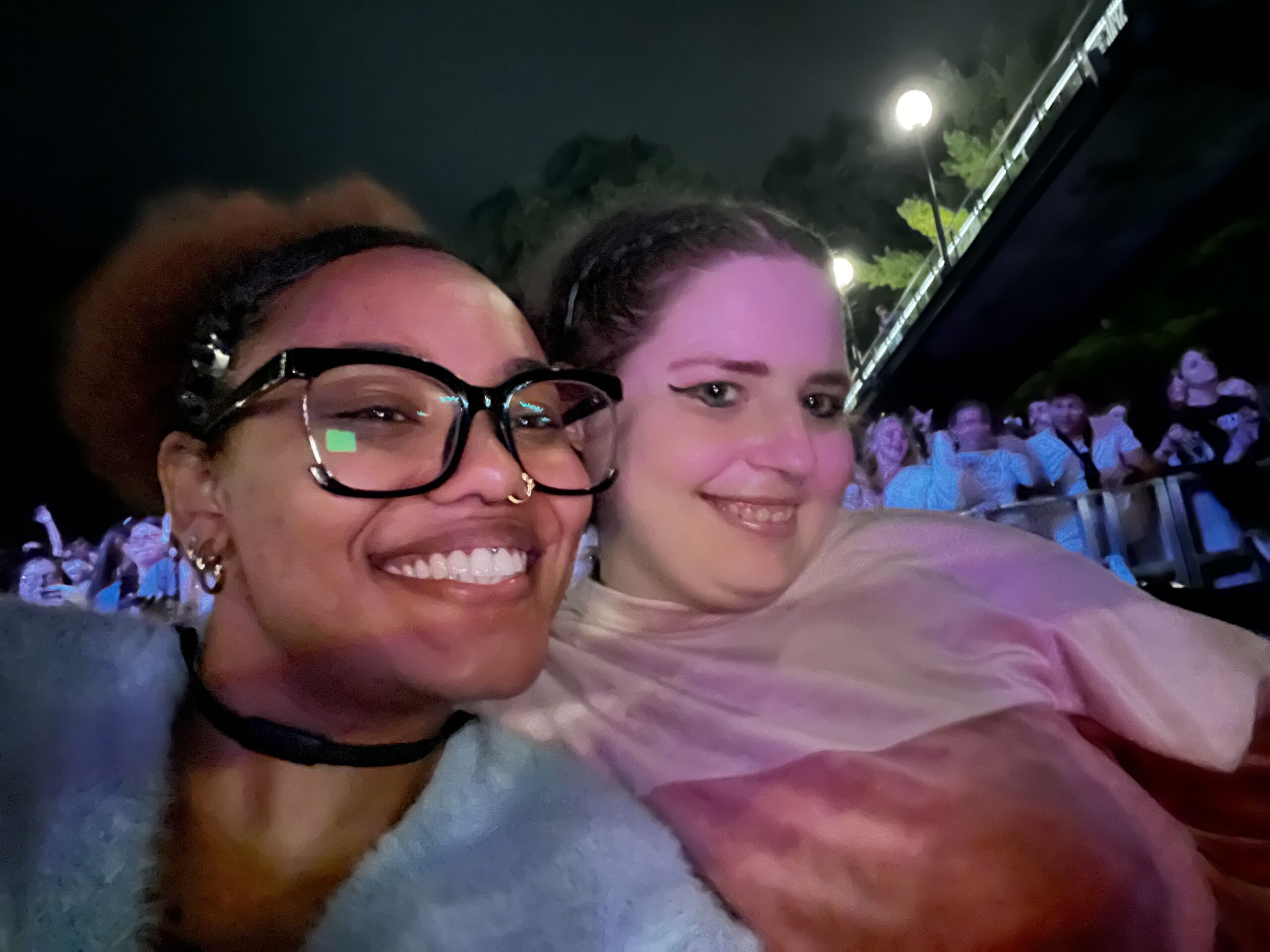 Pitbull Concert 2 Nighttime selfie of two friends posing close together at an outdoor event or concert venue. The person on the left wears black-framed glasses, a dark choker, and a fuzzy light blue sweater, smiling brightly at the camera. The person on the right wears winged eyeliner and a pink top, smiling softly. Pink and purple concert lighting illuminates their faces, with a crowd of people and venue lights visible in the blurred background.