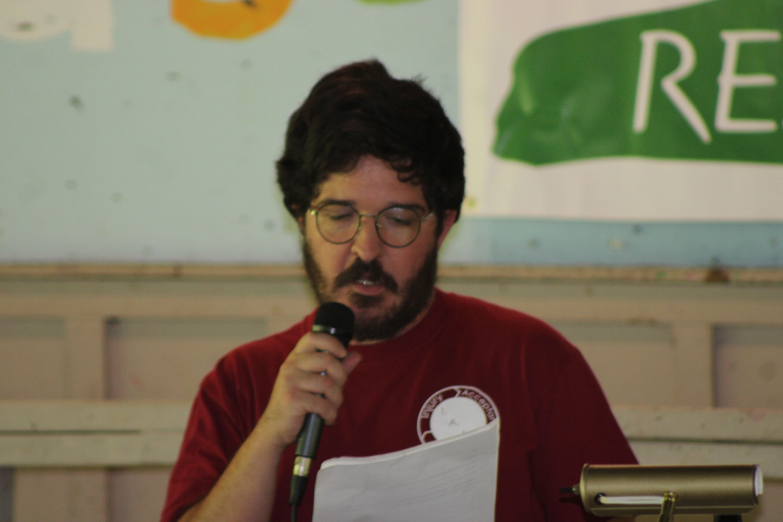 Brain Injury Awareness Picnic Speech A man with dark hair, a beard, and round glasses wearing a red t-shirt, standing indoors and reading papers aloud into a handheld microphone.