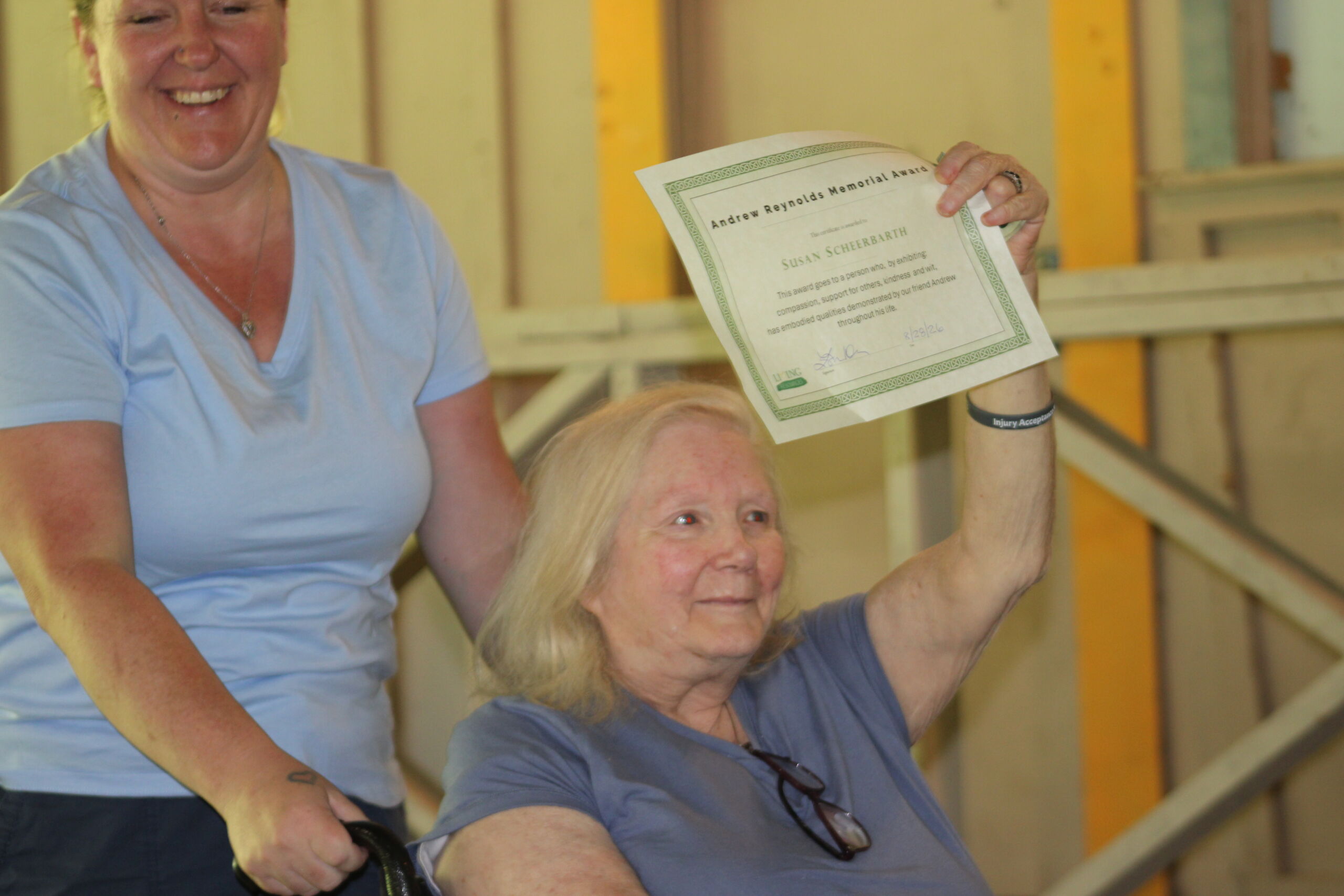 Brain Injury Awareness Day Awards An older blonde woman sitting in a wheelchair smiling and proudly holding up a certificate titled "Andrew Reynolds Memorial Award" presented to Susan S., while a smiling woman in a light blue t-shirt stands behind her.
