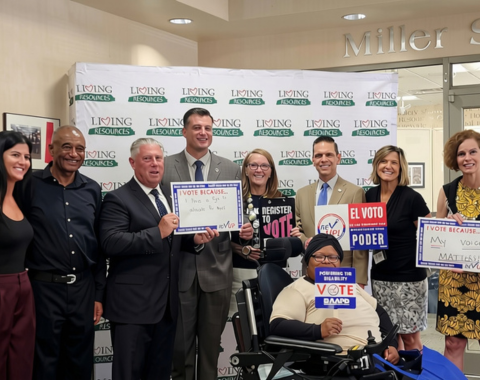 A group of people posing in front of a Living Resources backdrop during a voter registration drive event. Several individuals hold signs promoting voting rights, including ones that read "I Vote Because... I have a right to advocate for myself," "Register to Vote," "REV UP!," and "El Voto Es Poder." A woman in the front center uses a motorized wheelchair and holds a sign reading "Powering the Disability Vote."