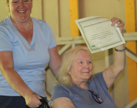 An older blonde woman sitting in a wheelchair smiling and proudly holding up a certificate titled "Andrew Reynolds Memorial Award" presented to Susan S., while a smiling woman in a light blue t-shirt stands behind her.