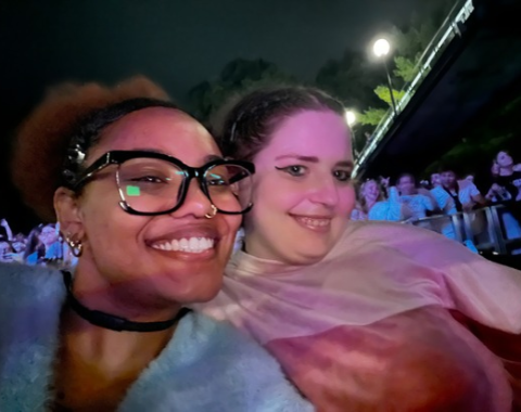 Nighttime selfie of two friends posing close together at an outdoor event or concert venue. The person on the left wears black-framed glasses, a dark choker, and a fuzzy light blue sweater, smiling brightly at the camera. The person on the right wears winged eyeliner and a pink top, smiling softly. Pink and purple concert lighting illuminates their faces, with a crowd of people and venue lights visible in the blurred background.