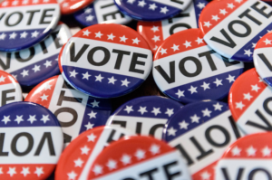 Vote Buttons A close-up photograph featuring a collection of round, patriotic campaign pins in red, white, and blue. Each button prominently reads "VOTE" in bold block lettering, decorated with white stars along the top or bottom edges.