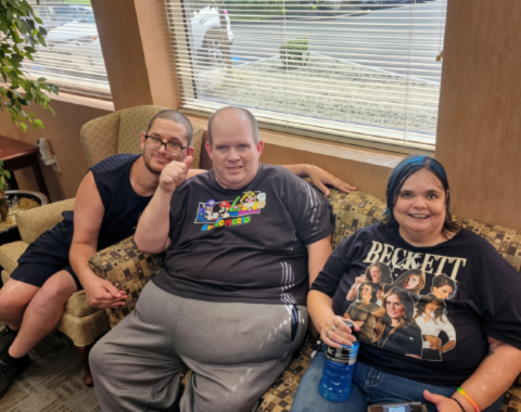 Voter Registration Drive 1 Three people sitting together on a pattern-upholstered couch inside an office waiting area near a window. The person on the left gives a thumbs-up, the person in the middle wears a Super Mario T-shirt and smiles, and the person on the right wears a graphic T-shirt with blue hair highlights while holding a sports drink bottle.