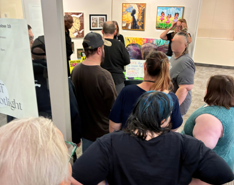 Voting Registration Line A group of people waiting in line inside an art gallery space during a voter registration event. Artwork hangs on the brightly lit walls in the background, and attendees gather around an information table set up in the room.