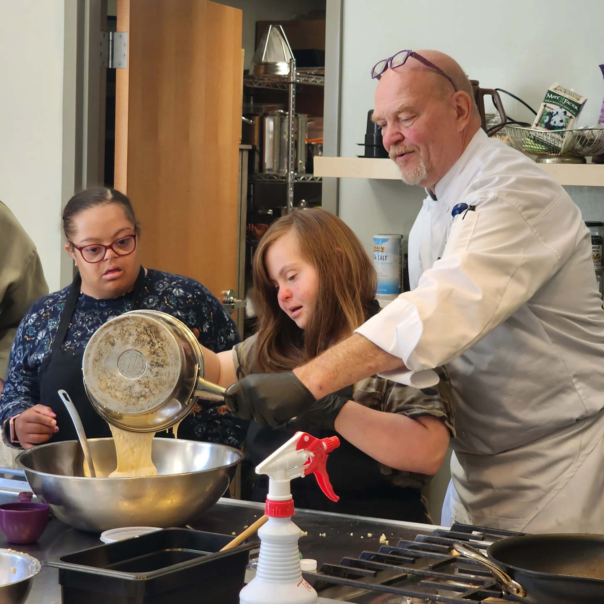 A male chef in a white uniform assists a participant wearing black gloves as she pours a warm sauce from a saucepan into a large mixing bowl, while another participant observes in the kitchen.