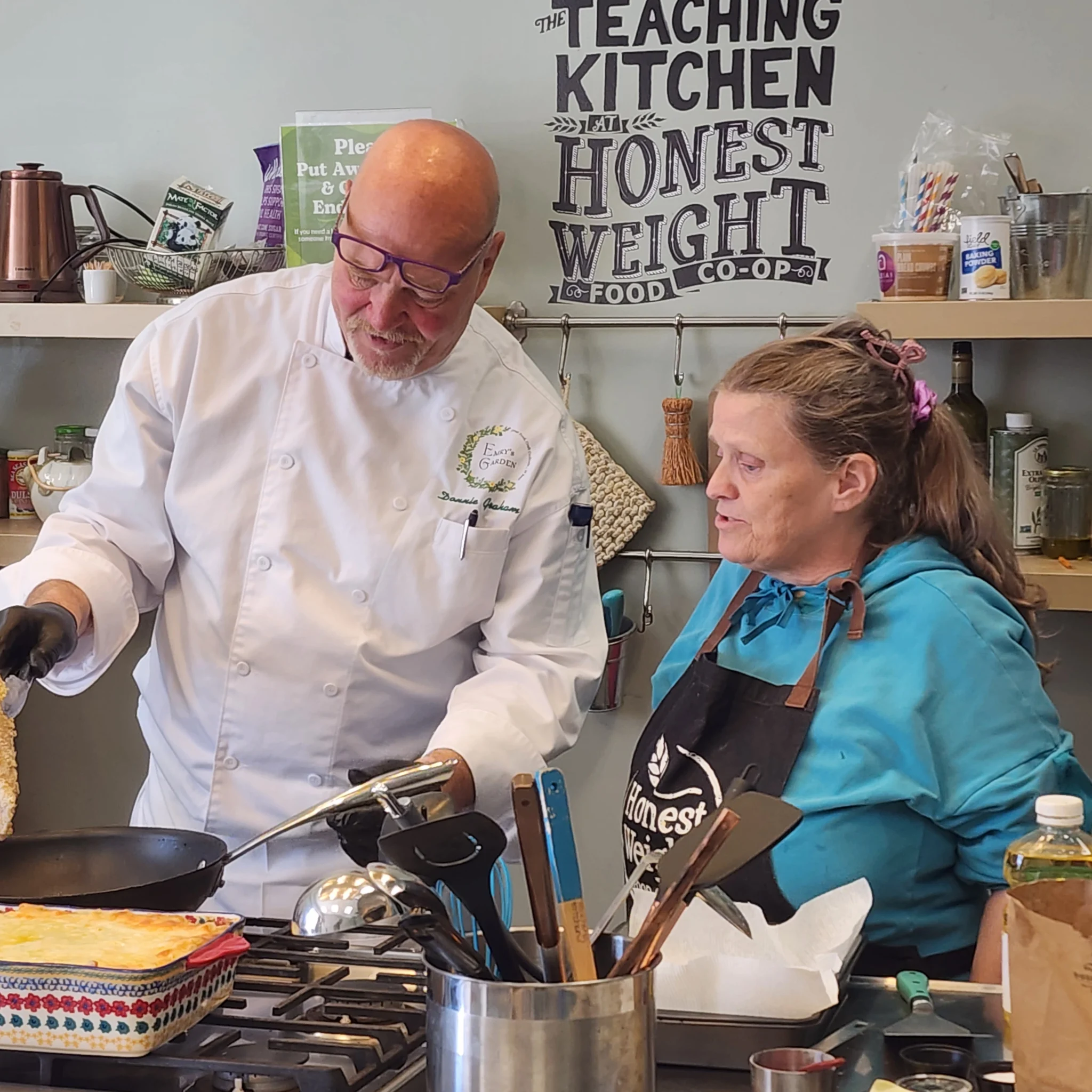 A chef in a white coat demonstrates a cooking technique in front of a wall sign that reads "The Teaching Kitchen at Honest Weight Food Co-op," as a woman in a blue hoodie and apron watches intently.