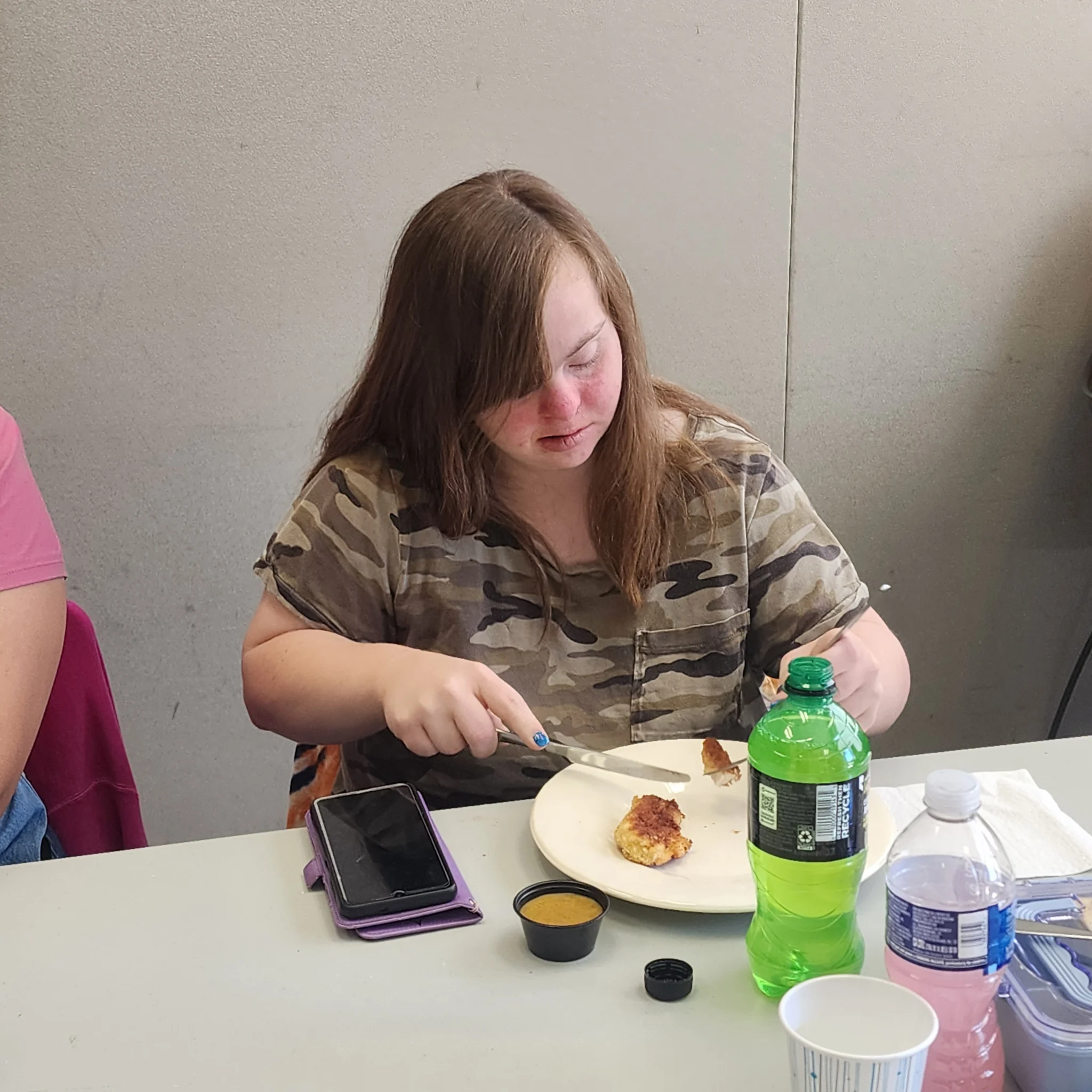 A young woman with brown hair in a camouflage shirt sits at a dining table, using a knife and fork to cut into a piece of crispy chicken on her plate alongside drinks and dipping sauce.