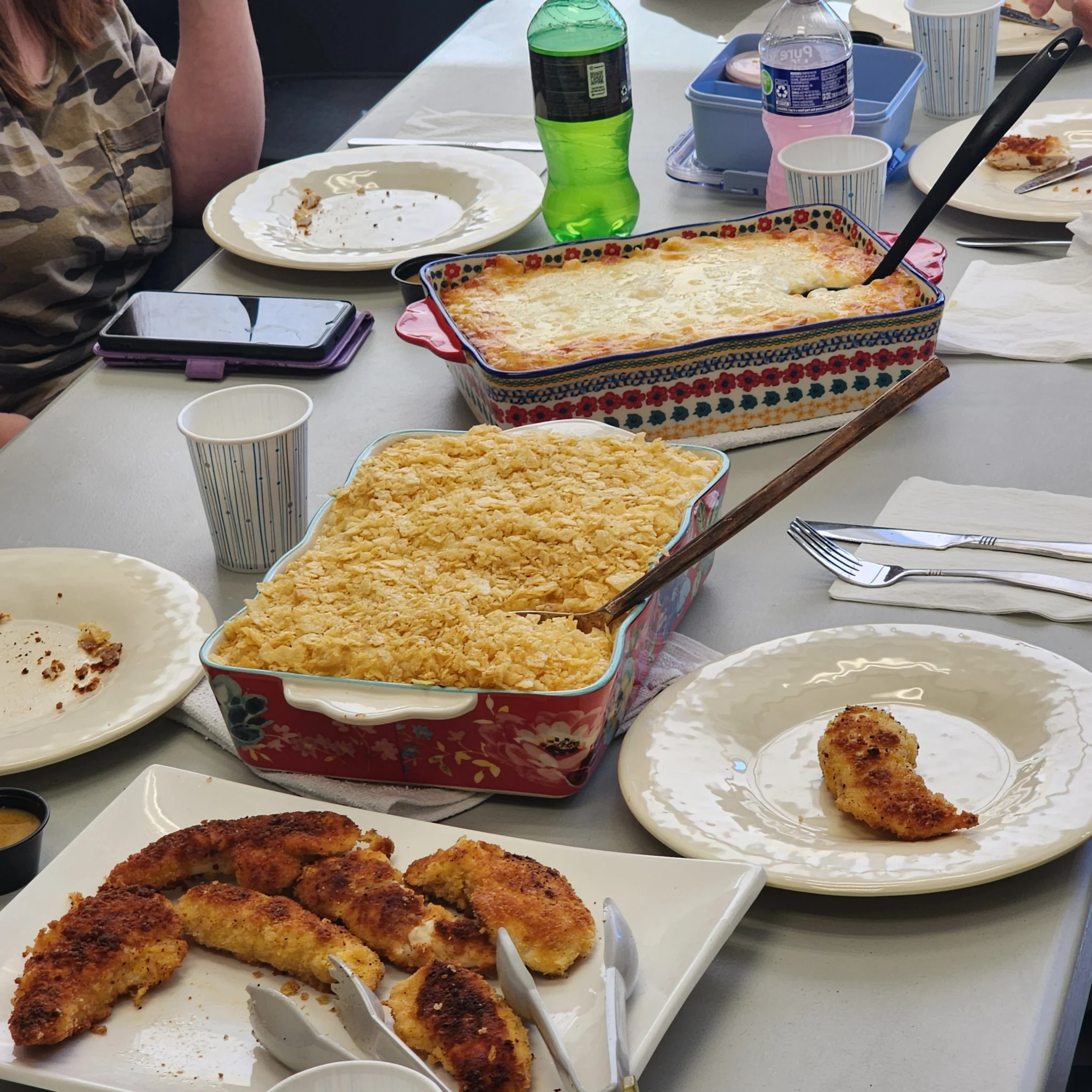 A dining table set with a family-style meal featuring a platter of golden-brown chicken tenders, two large baking dishes of casserole and macaroni, plates, and beverages.
