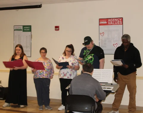 A group of five singers stands in a row holding binders and music sheets during a choir practice or performance. In the foreground, viewed from behind, a keyboardist plays an electronic keyboard set up on a stand with music scores. On the white wall behind them are posters titled "Agency Values" featuring word puzzles.