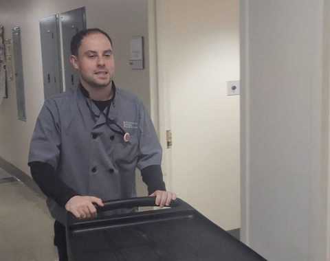Brendan smiling as he pushes a black utility cart down a brightly lit building hallway. He is dressed in a gray double-breasted chef-style uniform coat featuring a logo on the left chest, a pin attached to his lanyard, and a black long-sleeve shirt underneath.
