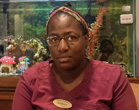 A healthcare worker named Denise wearing maroon scrub tops and a name badge reading "Teresian House - Denise, CNA." She wears glasses, hoop earrings, and a patterned headband in her styled hair, facing forward with a calm expression. Behind her is a illuminated fish tank filled with green aquatic plants, decorative mushrooms, and rock ornaments.