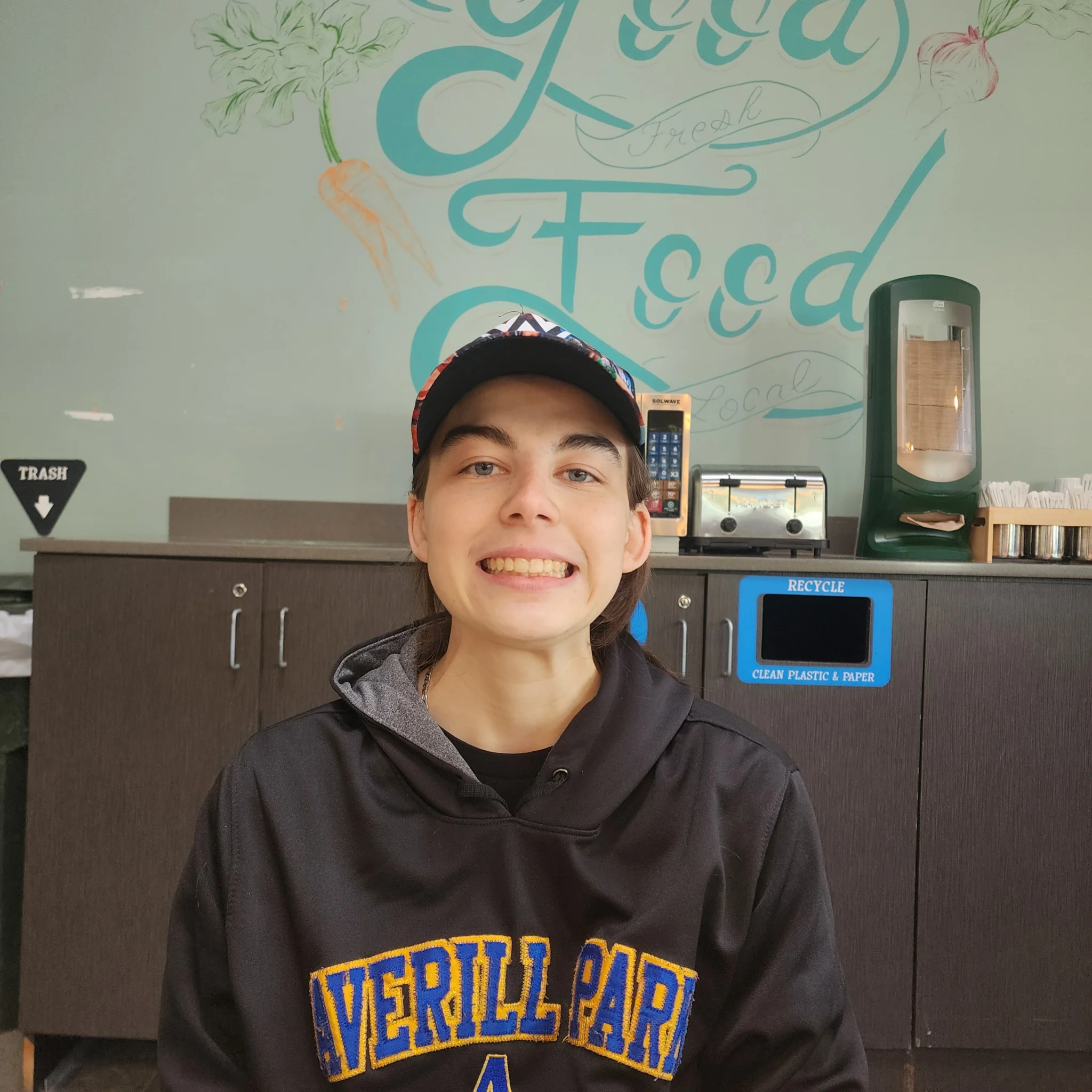 Honest Weight 1 A person with short dark hair and a baseball cap smiling at the camera, wearing a dark hoodie with yellow lettering. They are standing indoors in front of a counter with a green wall painted with the words "Good Food" and illustrated vegetables.