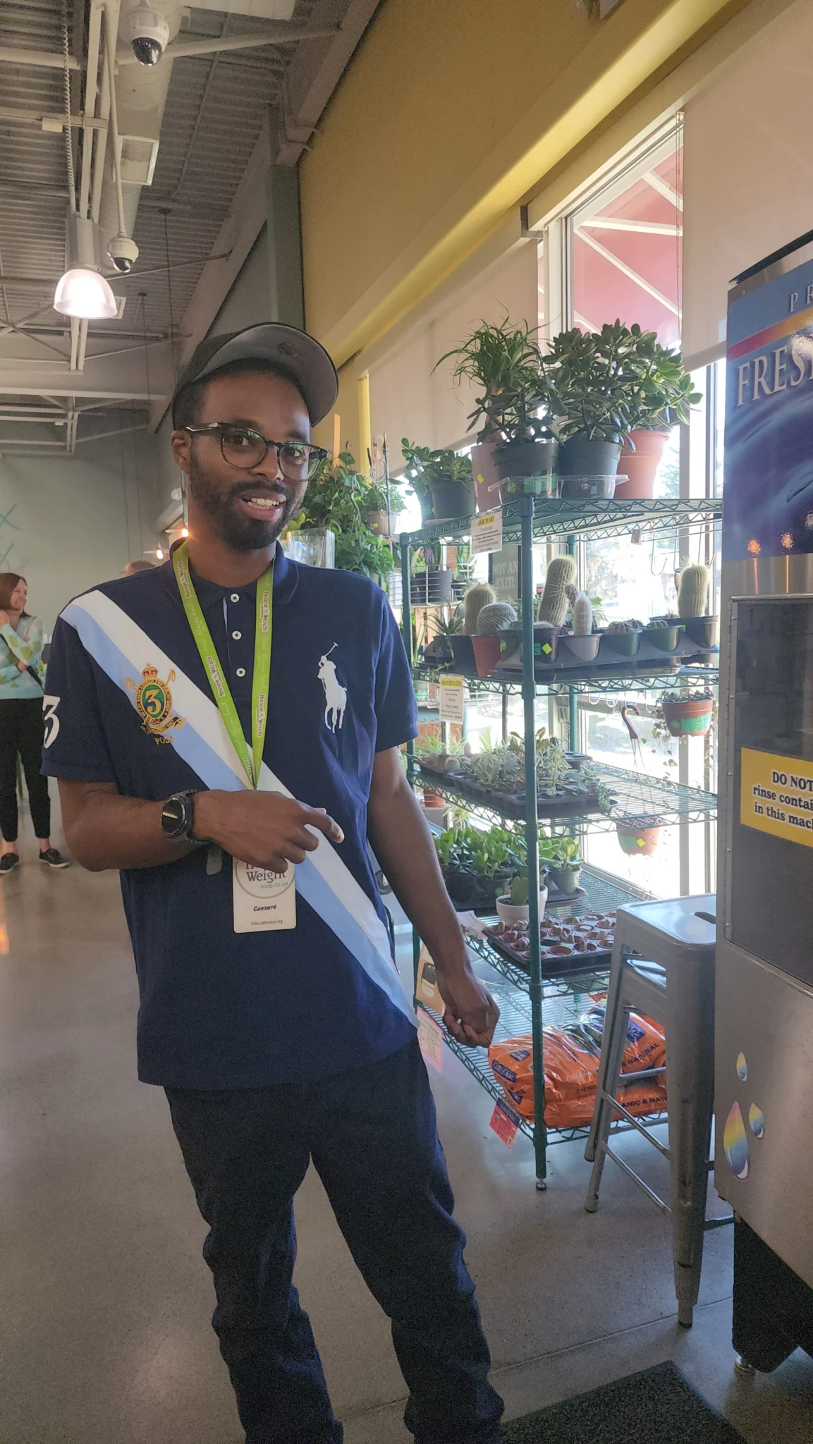 Honest Weight 2 A man with glasses, a beard, and a cap smiling while wearing a dark blue polo shirt with a light blue and white diagonal sash and a lanyard. He is standing indoors near a metal shelf holding potted plants and a large vending machine.