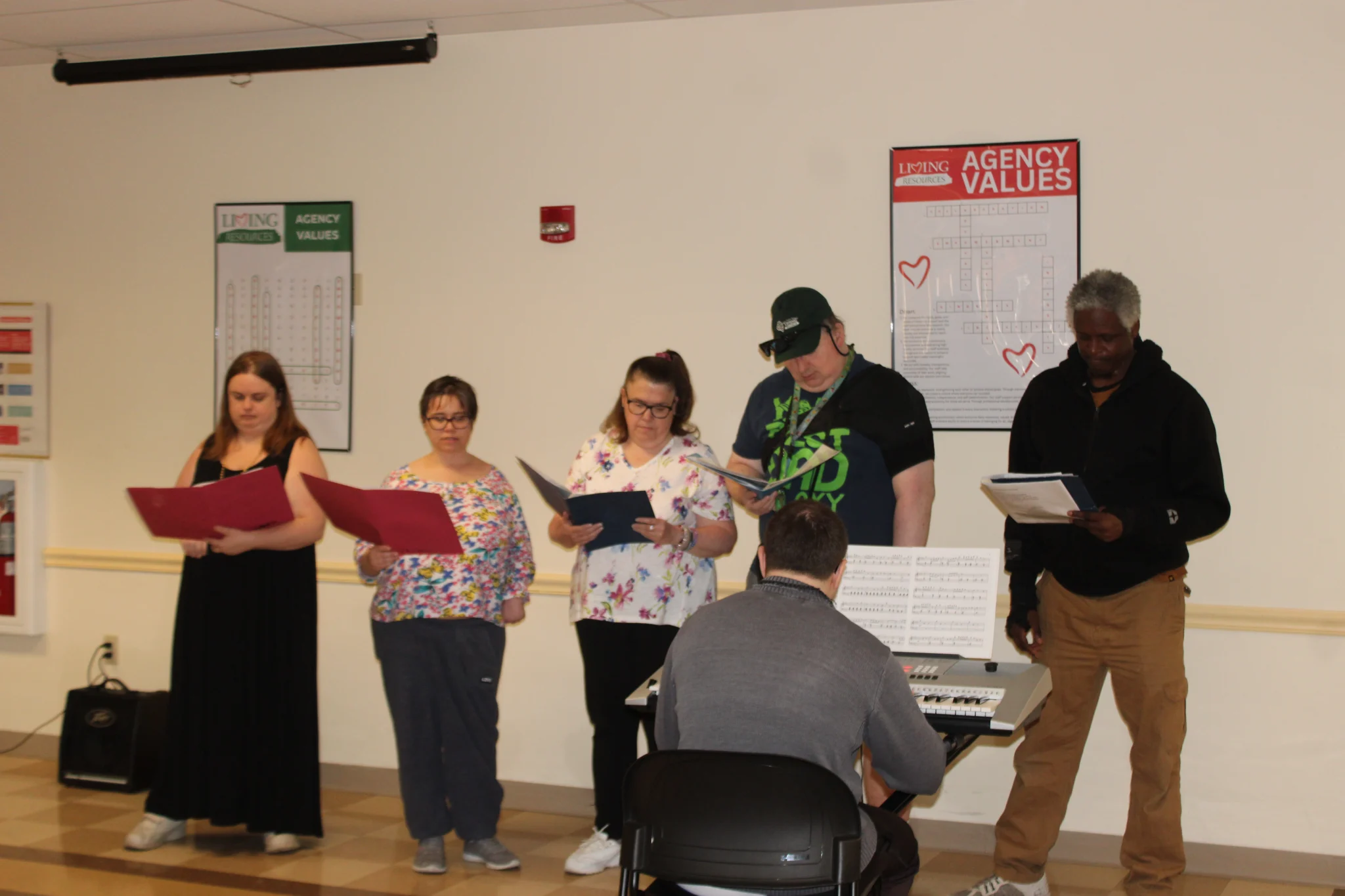 ABI Chorus A group of five singers stands in a row holding binders and music sheets during a choir practice or performance. In the foreground, viewed from behind, a keyboardist plays an electronic keyboard set up on a stand with music scores. On the white wall behind them are posters titled "Agency Values" featuring word puzzles.