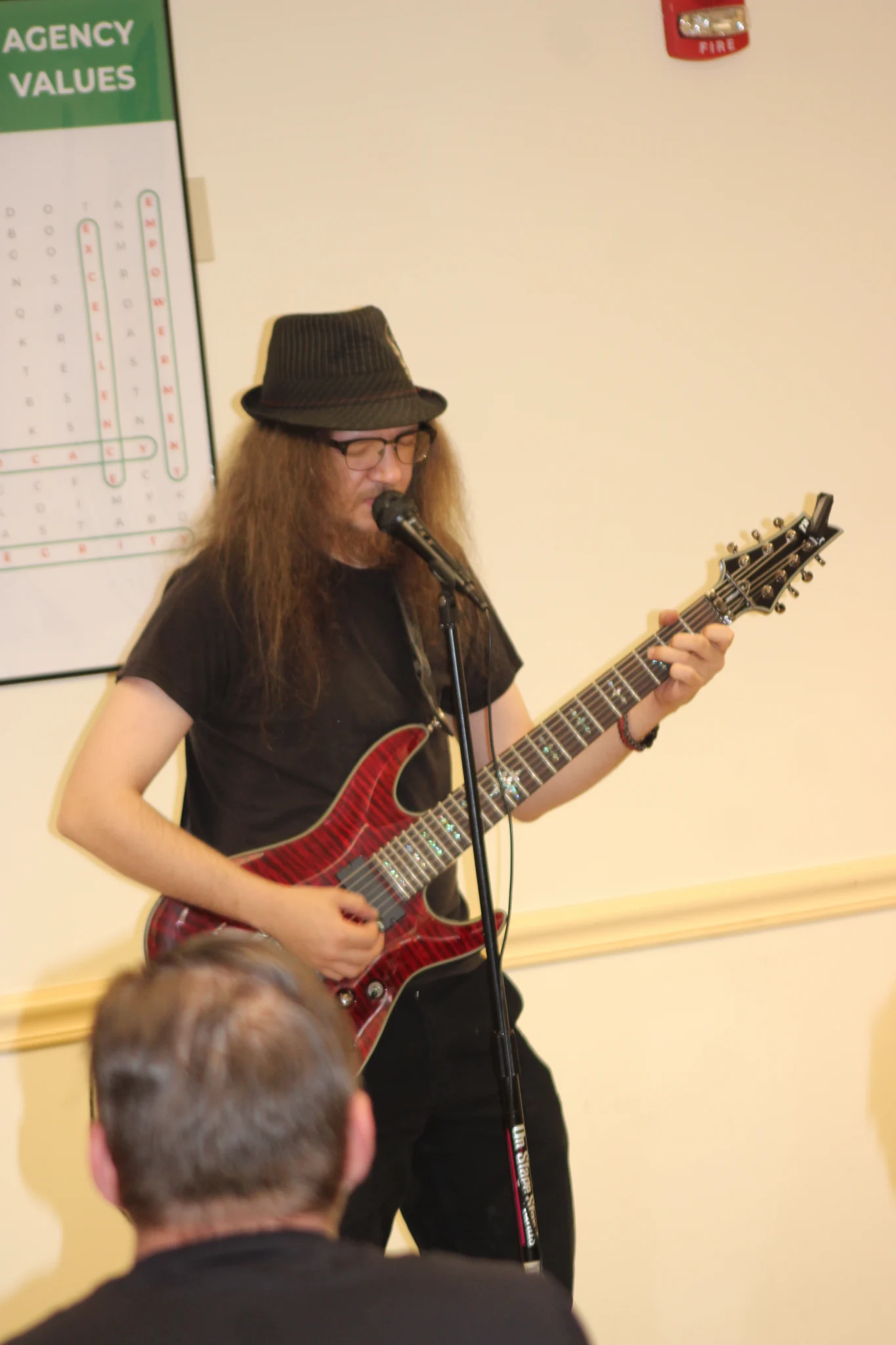 ABI Talent Show Guitar Performance A musician wearing a black hat, glasses, and a black t-shirt performs on stage, playing a red patterned electric guitar and singing into a microphone on a stand. The back of an audience member's head is visible in the lower foreground facing the performer.