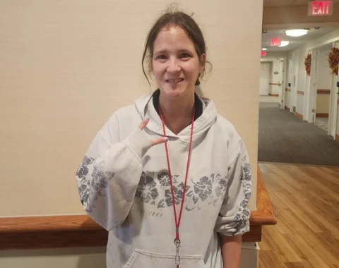 Medium close-up portrait of Kaylee, a woman with her hair pulled back, smiling at the camera. She is wearing a off-white hoodie featuring a floral pattern on the sleeves and chest, along with a red lanyard around her neck. She is making a hand gesture with her right hand while standing in a brightly lit indoor hallway.