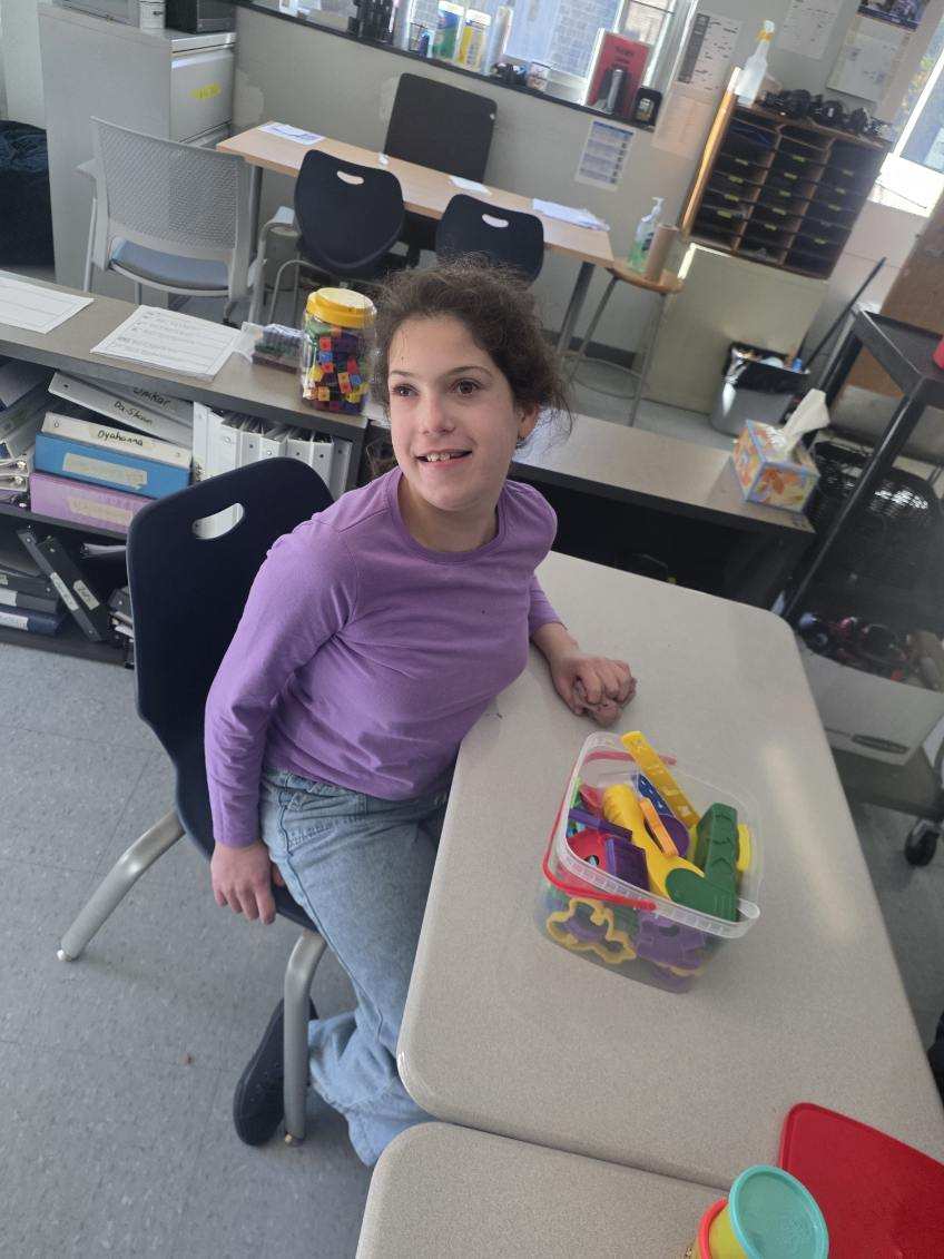 Girl with Play-Doh Tools A young girl in a purple long-sleeved shirt and blue jeans sits smiling at a classroom desk. Next to her on the desk is a clear container filled with colorful plastic dough tools and shape cutters.